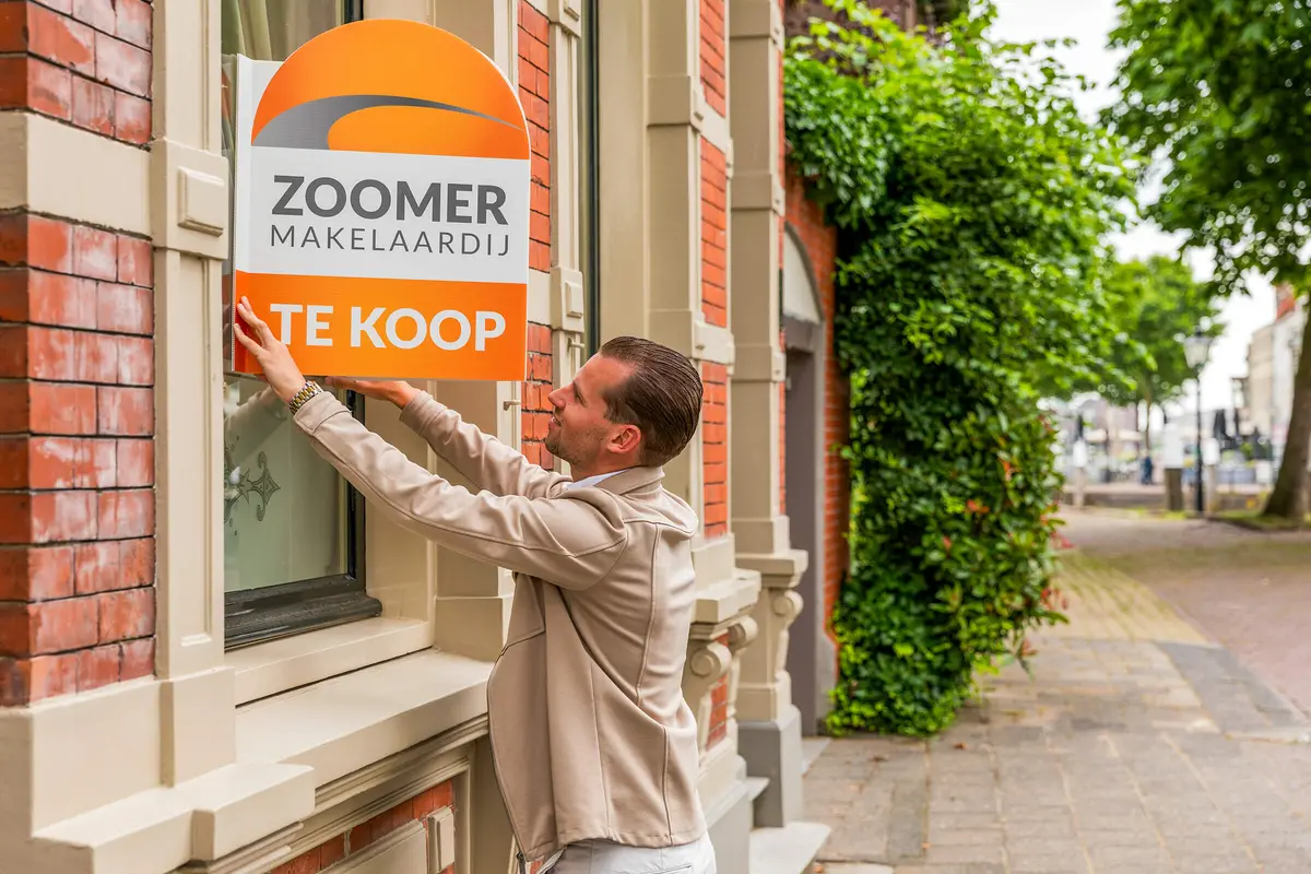 Makelaar van Zoomer hangt een te koop-bord op bij een woning